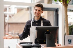 Smiling young man in apron standing at the cash register indoors