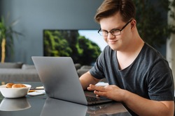 Smiling white young man with down syndrome using laptop computer at home sitting at the table