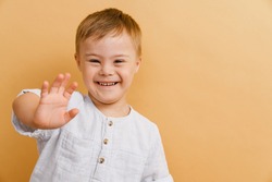 White boy with down syndrome smiling and gesturing at camera isolated over beige background
