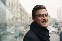 Portrait of young smiling european businessman looking at camera. Concept of modern successful man. Handsome stylish guy wearing scarf, coat and glasses. Blurred city background at cold day