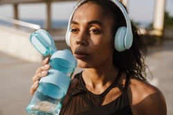 Black sportswoman listening music and drinking water while working out on parking outdoors