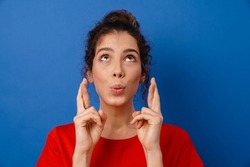 Young surprised woman holding fingers crossed for good luck isolated over blue background