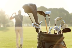 Selective focus of backpack with different putters with background of man playing golf on green lawn at sunny day. Concept of entertainment, recreation, leisure and hobby outdoors