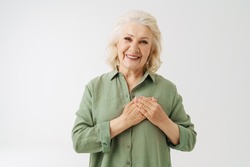 Grey senior woman in shirt smiling while holding her hands on chest isolated over white background