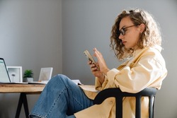 Young curly serious woman in glasses with phone sitting by table in cozy room at home