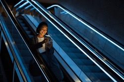 Young joyful asian woman wearing suit using mobile phone while standing on escalator indoors