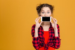 Portrait of a young woman covering her mouth with blank smartphone screen over yellow background