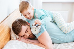 Cute little son holding alarm clock near sleeping father ear at home