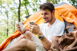 Cheerful young man tourist sitting and charging battery of mobile phone in touristic tent