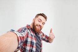 Close up portrait of a happy casual man taking selfie and showing thumbs up gesture over white background