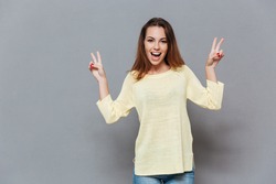 Portrait of an excited young woman in sweater showing peace signs with hands isolated on the gray background