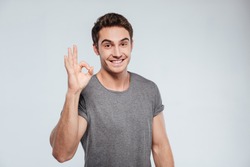 Portrait of a cheerful young man showing okay gesture isolated on the gray background