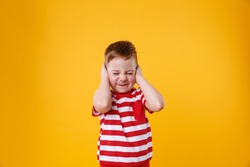 Portrait of an angry unhappy irritated little boy covering ears isolated over orange background