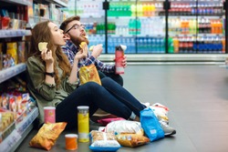 Portrait of a young funny hungry couple sitting on the supermarket floor and eating junk food