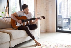 Pretty young man playing guitar while sitting on sofa in light living room