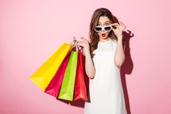 Image of a shocked young brunette lady in white summer dress wearing sunglasses posing with shopping bags and looking at camera over pink background.