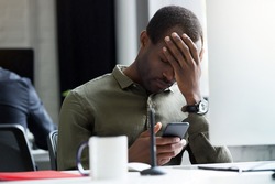 Upset young african man reading message on his mobile phone while sitting at his desk