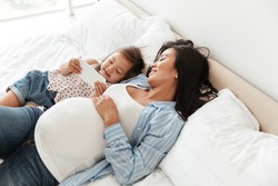 Happy pregnant woman and her little daughter using mobile phone while relaxing together in bed at home