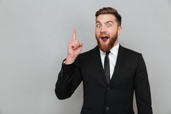 Excited young bearded businessman having an idea and pointing finger up at copy space isolated over gray background