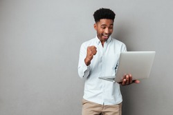 Excited happy afro american man looking at laptop computer screen and celebrating the win isolated over gray background