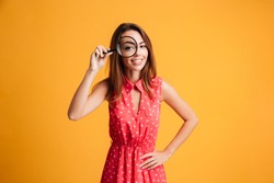 Close-up portrait of cheerful pretty woman in red dress looking at camera through magnifying glass, isolated over yellow background