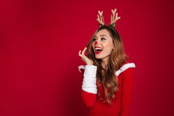 Portrait of an cheerful happy girl wearing christmas deer costume while standing and looking away at copy space isolated over red background