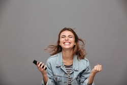Close-up portrait of beautiful screaming young girl holding smartphone showing winner gesture, isolated on gray background