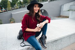 Portrait of a cheerful happy asian girl wearing hat and sweater holding coffee cup while sitting on a city street