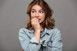 Close up portrait of a worried teenage girl in denim jacket biting her nails and looking away isolated over gray background