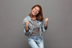 Portrait of a joyful happy teenage girl dressed in denim jacket celebrating success while dancing isolated over gray background
