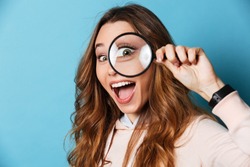 Close up portrait of a cheerful pretty girl looking through magnifying glass isolated over blue background