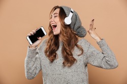 Portrait of emotional young woman in gray woolen hat holding her smartphone like microphone and singing, isolated on beige background