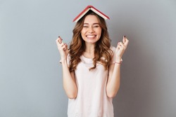 Portrait of a happy girl holding book on her head with crossed fingers for good luck isolated over gray wall background