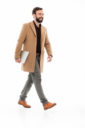 Full length portrait of a confident bearded guy dressed in coat holding laptop computer while walking isolated over white background