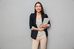 Pleased asian business woman holding laptop computer and looking at the camera over gray background