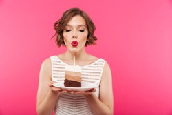 Portrait of a pretty girl holding plate with a piece of birthday cake and blowing a candle isolated over pink background