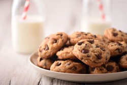 Pile of Homemade Chocolate Chip Cookies on a plate, on a wood white table, with milk bottles on the background
