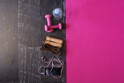 Top view image of jumping rope, smart phone, pink yoga mat and weights with a bottle of water on black wooden floor background. Equipment for fitness. Concept healthy lifestyle. Lots of copy space.