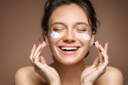Laughing girl applying moisturizing cream on her face. Photo of young girl with flawless skin on brown background. Skin care and beauty concept