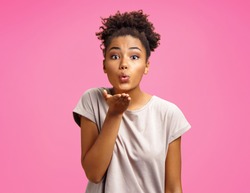 Pretty girl makes air kiss into camera, expresses her love. Photo of african american girl wears casual outfit on pink background. Emotions and pleasant feelings concept.