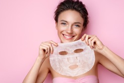 Smiling woman holds facial sheet mask. Photo of attractive woman with perfect skin on pink background. Beauty & Skin care concept