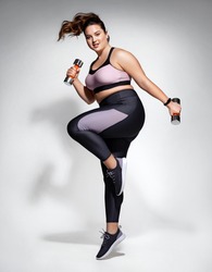 Sporty girl jumping with dumbbells. Photo of model with curvy figure in fashionable sportswear on grey background. Dynamic movement. Side view. Sports motivation and healthy lifestyle