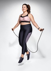 Sporty girl with skipping rope in motion. Photo of model with curvy figure in fashionable sportswear on grey background. Dynamic movement. Sports motivation and healthy lifestyle