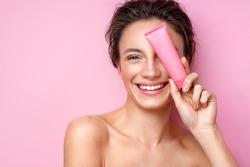 Smiling woman holds tube with cosmetic cream. Photo of attractive woman with perfect makeup on pink background. Beauty concept