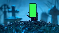 Zombie hand rising up smartphone with green screen out of grave. Holiday event halloween concept.