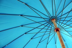 Under a blue parasol showing the details of the metal spokes. The umbrella is providing sun block yet projecting a vivid aqua blue background color.