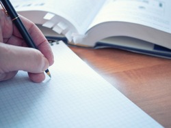 left handed student with pen preparing for exams, writing notes at textbook on wooden desk. opened encyclopedia dictionary with bookmarks on background out of focus. closeup image