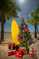 The decorated Christmas tree with flip flops and sunglasses together with presents and surf board on the beach, the Christmas vacation concept of travel
