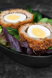 Scotch egg halved on lettuce leaves in a black dish.  On a stone background