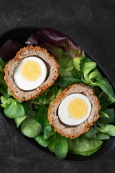Scotch egg halved on lettuce leaves in a black dish.  On a stone background